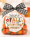 A clear cellophane gift bag filled with small orange pumpkin-shaped candies, tied with a black-and-white gingham bow. A round printed label on the front reads, 'Thank you for helping me FALL in love with Learning' with autumn-themed decorations.