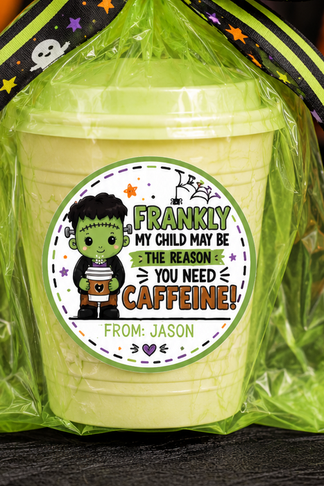 A Halloween-themed candy bucket gift bag filled with pale yellow candy, wrapped in bright green cellophane and tied with a black and green striped bow featuring ghosts and stars. The front has a circular custom label that reads 'FRANKLY MY CHILD MAY BE A CAFFEINE! FROM: JASON' with a cartoon Frankenstein character.