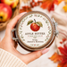 A hand holds a glass jar of apple butter with a decorative lid label featuring an apple illustration and the text 'APPLE BUTTER made with love from: Jamie's Kitchen.' The scene is styled with apples in a basket and autumn leaves on a light surface.