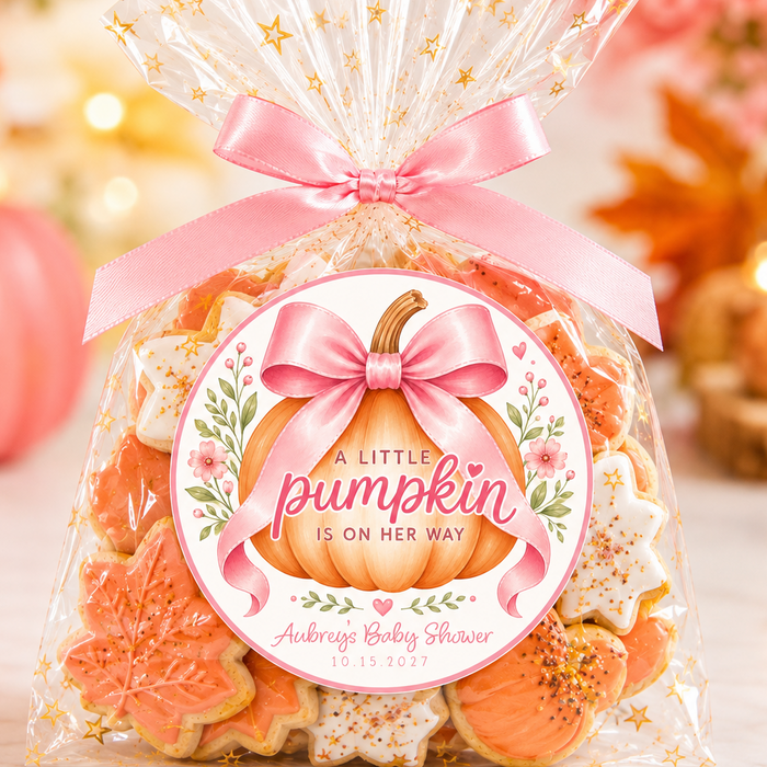 A clear cellophane gift bag filled with assorted fall-themed cookies, tied with a pink satin ribbon and topped with a clear bag featuring gold stars. The front has a round printed label reading 'A Little Pumpkin Is On Her Way' with a pumpkin illustration and personalized text for a baby shower.