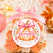 A clear cellophane gift bag filled with assorted fall-themed cookies, tied with a pink satin ribbon and topped with a clear bag featuring gold stars. The front has a round printed label reading 'A Little Pumpkin Is On Her Way' with a pumpkin illustration and personalized text for a baby shower.