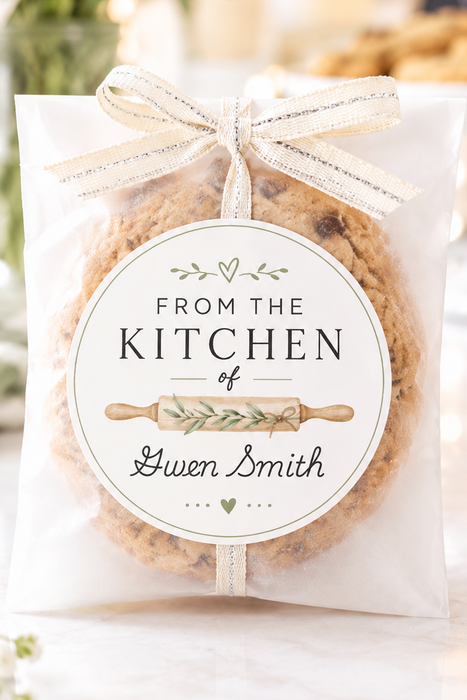 A round chocolate chip cookie sealed in a clear gift bag with a white ribbon bow and a personalized round label reading 'FROM THE KITCHEN of Gwen Smith,' set on a light tabletop with blurred flowers in the background.