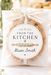 A round chocolate chip cookie sealed in a clear gift bag with a white ribbon bow and a personalized round label reading 'FROM THE KITCHEN of Gwen Smith,' set on a light tabletop with blurred flowers in the background.