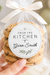 A hand holds a clear cellophane bag of cookies tied with a cream ribbon, featuring a round printed label that reads 'FROM THE KITCHEN of Gwen Smith.' The background is softly blurred with white flowers, balloons, and a cupcake, suggesting a wedding or celebratory setting.