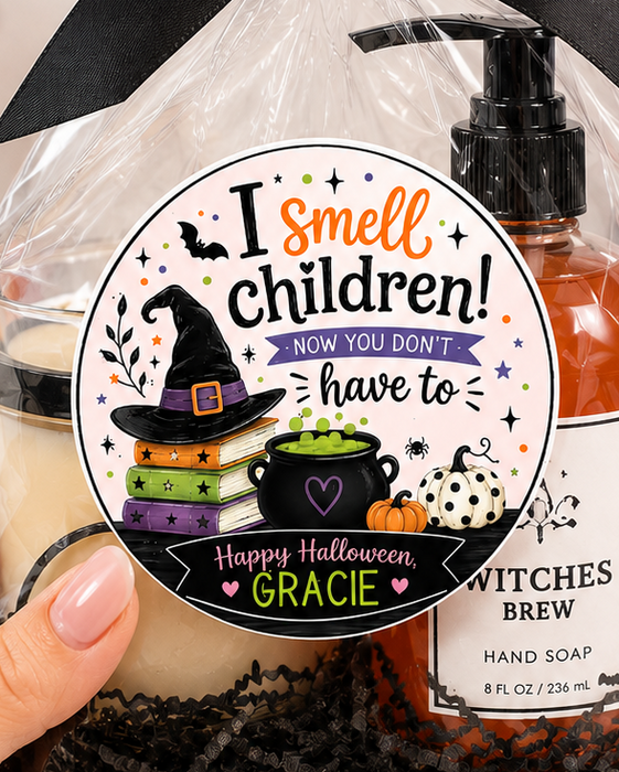 A hand holds a clear gift bag filled with Halloween-themed items, including a black ribbon bow, a candle, a bottle of Witch's Brew hand soap, and black shredded paper filler. The bag has a round sticker that reads 'I smell children! HOW YOU DON'T.