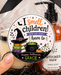 A hand holds a clear gift bag filled with Halloween-themed items, including a black ribbon bow, a candle, a bottle of Witch's Brew hand soap, and black shredded paper filler. The bag has a round sticker that reads 'I smell children! HOW YOU DON'T.