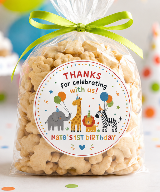 A clear cellophane bag filled with popcorn and tied with a bright green ribbon, featuring a round safari-themed 'Thanks for celebrating with us!' label. The bag is photographed on a party table with balloons, bunting, cupcakes, and a party hat in the background.