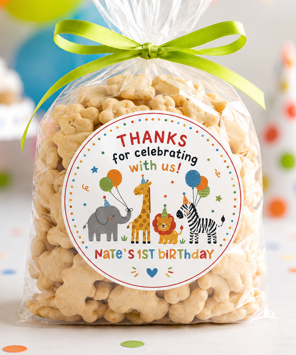 A clear cellophane bag filled with popcorn and tied with a bright green ribbon, featuring a round safari-themed 'Thanks for celebrating with us!' label. The bag is photographed on a party table with balloons, bunting, cupcakes, and a party hat in the background.