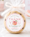 A round chocolate chip cookie wrapped in clear cellophane and tied with a white ribbon, featuring a personalized peach-themed thank you label that reads 'Thank You for showering our little peach Amber's baby shower.' The photo is set on a light marble surface with a soft pink floral background.