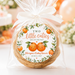 A round cookie wrapped in clear cellophane and tied with a white ribbon, featuring a round printed label that reads 'Two little cuties are on the way' with orange illustrations and the text 'Megan's Baby Shower June 8, 2027.' The background is softly blurred with warm orange and cream tones.