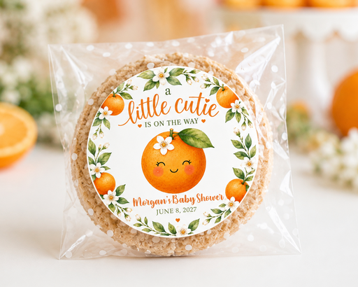 A round baked cookie or crisp in a clear cellophane bag with a printed orange-themed label reading 'a little catie is on the way' and 'Morgan's Baby Shower,' photographed on a bright white surface with blurred oranges and flowers in the background.