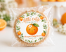 A round baked cookie or crisp in a clear cellophane bag with a printed orange-themed label reading 'a little catie is on the way' and 'Morgan's Baby Shower,' photographed on a bright white surface with blurred oranges and flowers in the background.
