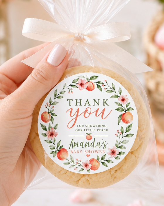 A hand holds a round thank-you cookie in a clear cellophane bag tied with a white ribbon, featuring a floral 'Thank You' label with peach illustrations and the text 'Amanda's Baby Shower.' The background is softly blurred with pink flowers and a candle.