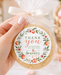 A hand holds a round thank-you cookie in a clear cellophane bag tied with a white ribbon, featuring a floral 'Thank You' label with peach illustrations and the text 'Amanda's Baby Shower.' The background is softly blurred with pink flowers and a candle.