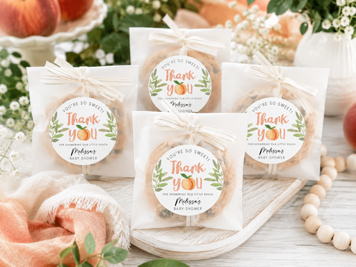 Four packaged baby shower cookies on a wooden tray with a peach and white floral background. Each cookie is in a clear bag tied with a ribbon, with a round printed label reading 'You're So Sweet Thank You' and personalized text.