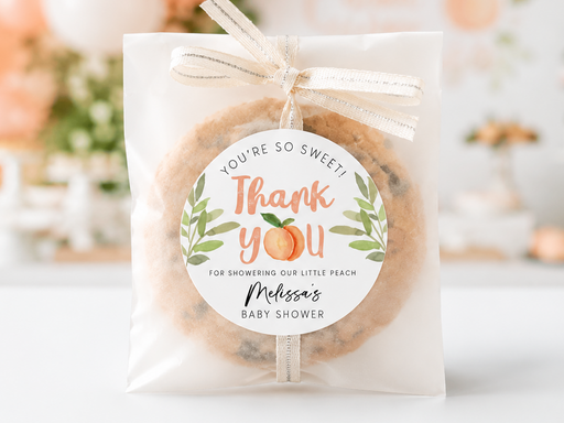 A clear plastic favor bag tied with a cream ribbon, containing a round chocolate chip cookie and featuring a circular printed thank-you label. The bag is centered on a white surface with a softly blurred peach and white party background.