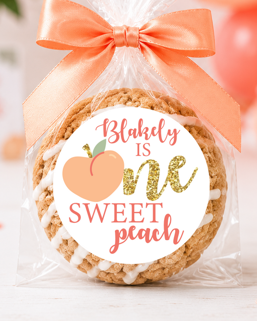 A round baked cookie wrapped in clear cellophane and tied with a peach satin bow, featuring a round white label that reads 'Blahely is one sweet peach' with a peach illustration. The scene is styled with peach balloons, flowers, confetti, and green leaves on a white surface.