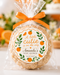 An orange-themed baby shower favor in a clear cellophane bag tied with an orange ribbon, featuring a round printed label that reads 'A little cutie is on the way Amanda's Baby Shower 11.25.