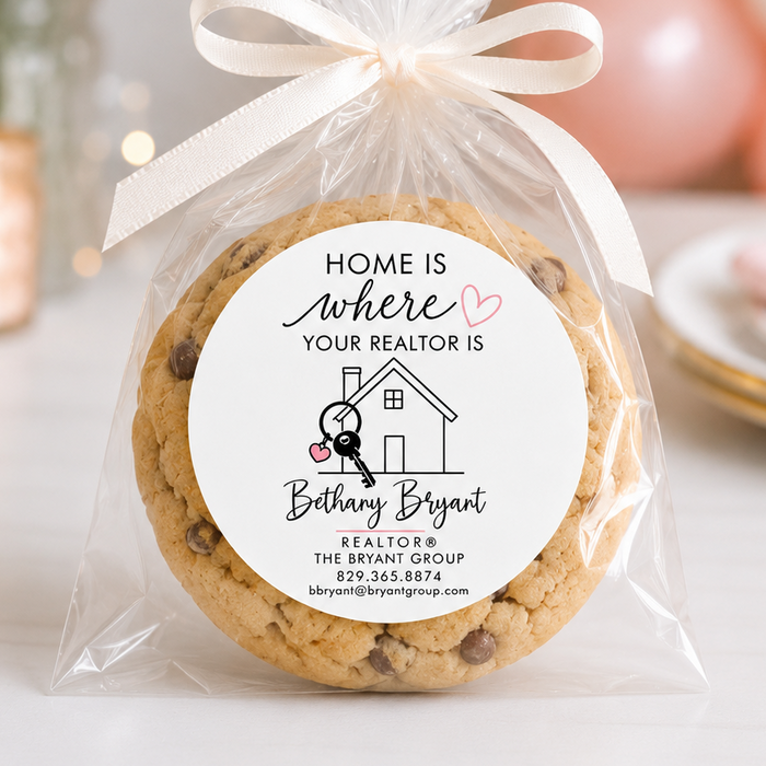 A round chocolate chip cookie wrapped in a clear cellophane bag tied with a cream ribbon, featuring a custom printed round label that reads 'HOME is where YOUR REALTOR IS' and the name 'Bethany Bryant'. The background is a softly blurred pink-themed party setting with balloons and plates.