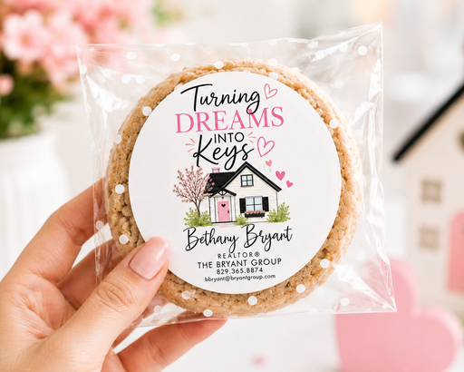 A hand holds a clear plastic bag containing a round custom cookie with a printed white label that reads 'Turning Dreams Into Keys Bethany Bryant' with a small house illustration. The background is softly blurred with pink flowers and a pink heart-shaped ornament.