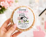 A hand holds a clear plastic bag containing a round custom cookie with a printed white label that reads 'Turning Dreams Into Keys Bethany Bryant' with a small house illustration. The background is softly blurred with pink flowers and a pink heart-shaped ornament.