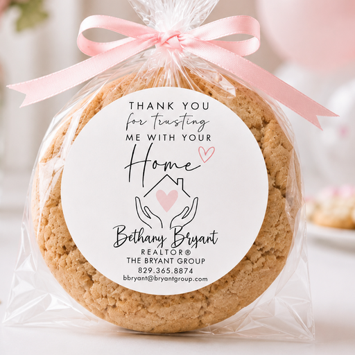 A round cookie wrapped in clear cellophane and tied with a pink ribbon, featuring a printed white label that reads 'Thank you for trusting me with your Home' and the name Bethany Bryant. The background is softly blurred with pink balloons and flowers.