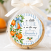 A round cookie wrapped in clear cellophane and tied with a cream ribbon, featuring a round printed label that reads 'a little CUTIE is on the way' with orange and blue floral artwork. The background is softly blurred with oranges and a cake.