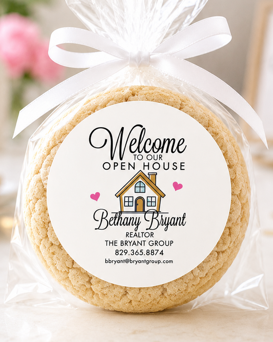 A round baked cookie wrapped in clear cellophane and tied with a white ribbon, featuring a printed circular label that reads 'Welcome to Our Open House Bethany Bryant' with a small house graphic. The scene is softly lit on a light surface with blurred decorative background elements including balloons and a house-shaped light.