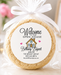 A round baked cookie wrapped in clear cellophane and tied with a white ribbon, featuring a printed circular label that reads 'Welcome to Our Open House Bethany Bryant' with a small house graphic. The scene is softly lit on a light surface with blurred decorative background elements including balloons and a house-shaped light.