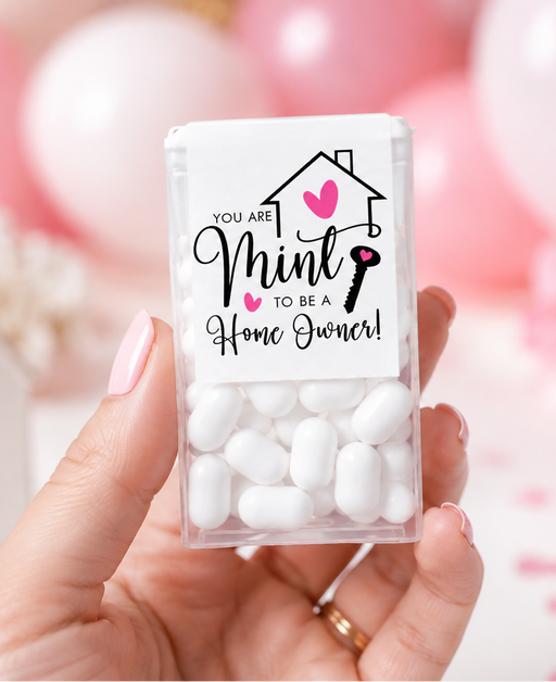 A hand holds a clear plastic jar of white candy-coated mint candies with a custom printed white label that reads 'YOU ARE Mint TO BE A Home Owner!' in black and pink text. The background is a soft-focus pink party setting with balloons, a small house decoration, and pink cups.