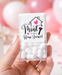 A hand holds a clear plastic jar of white candy-coated mint candies with a custom printed white label that reads 'YOU ARE Mint TO BE A Home Owner!' in black and pink text. The background is a soft-focus pink party setting with balloons, a small house decoration, and pink cups.