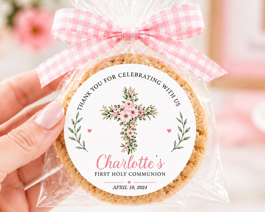 A hand holds a clear cellophane-wrapped round cookie or biscuit with a pink gingham bow. The front has a round printed label with a floral cross, the text 'Thank you for celebrating with us', the name 'Charlotte's', and 'First Holy Communion' with a date.