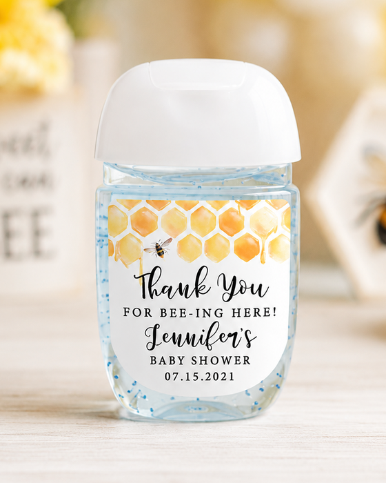 A small hand sanitizer bottle with a white cap and a honeybee-themed thank you label, set on a light wooden surface with a blurred yellow and white floral background.
