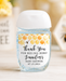 A small hand sanitizer bottle with a white cap and a honeybee-themed thank you label, set on a light wooden surface with a blurred yellow and white floral background.