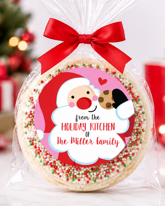 A round holiday treat cookie wrapped in clear cellophane and tied with a red ribbon, featuring a personalized Santa label that reads 'from the Holiday Kitchen of the Miller Family.' The background shows a softly blurred Christmas tree, ornaments, and red gift boxes.