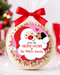 A round holiday treat cookie wrapped in clear cellophane and tied with a red ribbon, featuring a personalized Santa label that reads 'from the Holiday Kitchen of the Miller Family.' The background shows a softly blurred Christmas tree, ornaments, and red gift boxes.