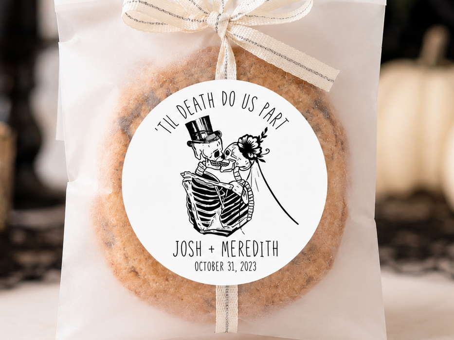 A round sugar cookie wrapped in a clear cellophane bag tied with a cream ribbon, featuring a circular printed label with a skeleton and rose illustration and the text 'I'D U DEATH DO US PART JOSH + MEREDITH OCTOBER 31, 2021.' The package is photographed on a tabletop with candles, black lace, and pumpkins in the background.