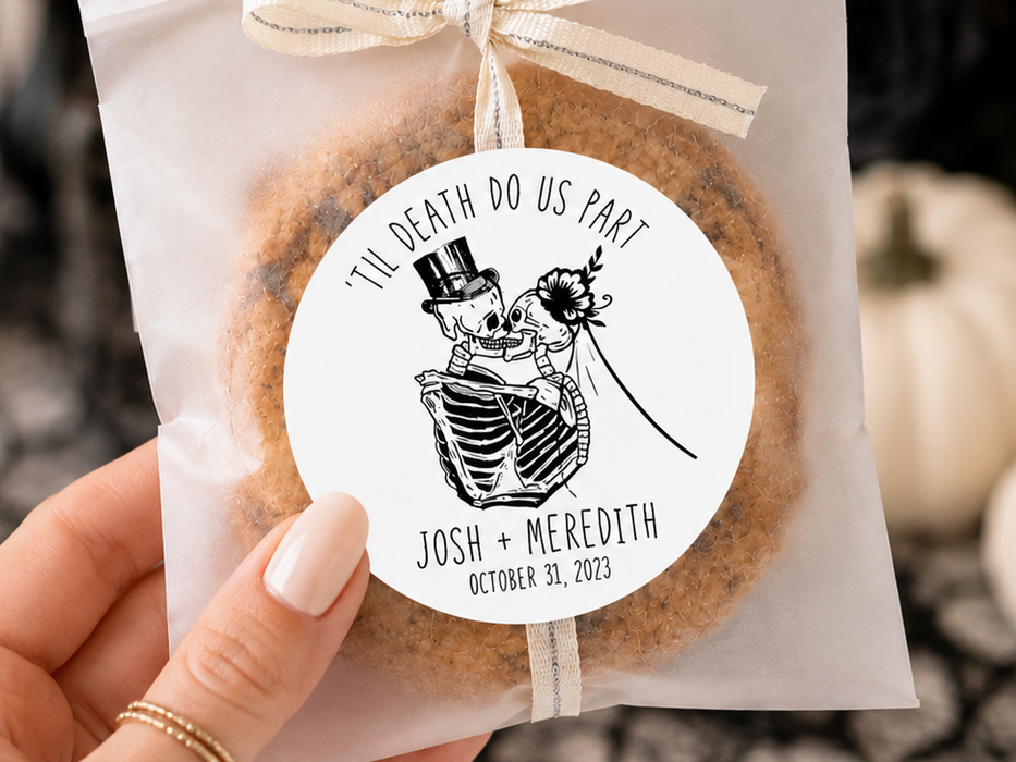 A hand holds a clear bag containing a round decorated cookie or biscuit with a cream ribbon bow and a white circular label. The label has a black skeleton wedding illustration and the text “I’m Death Do Us Part” with names and date.