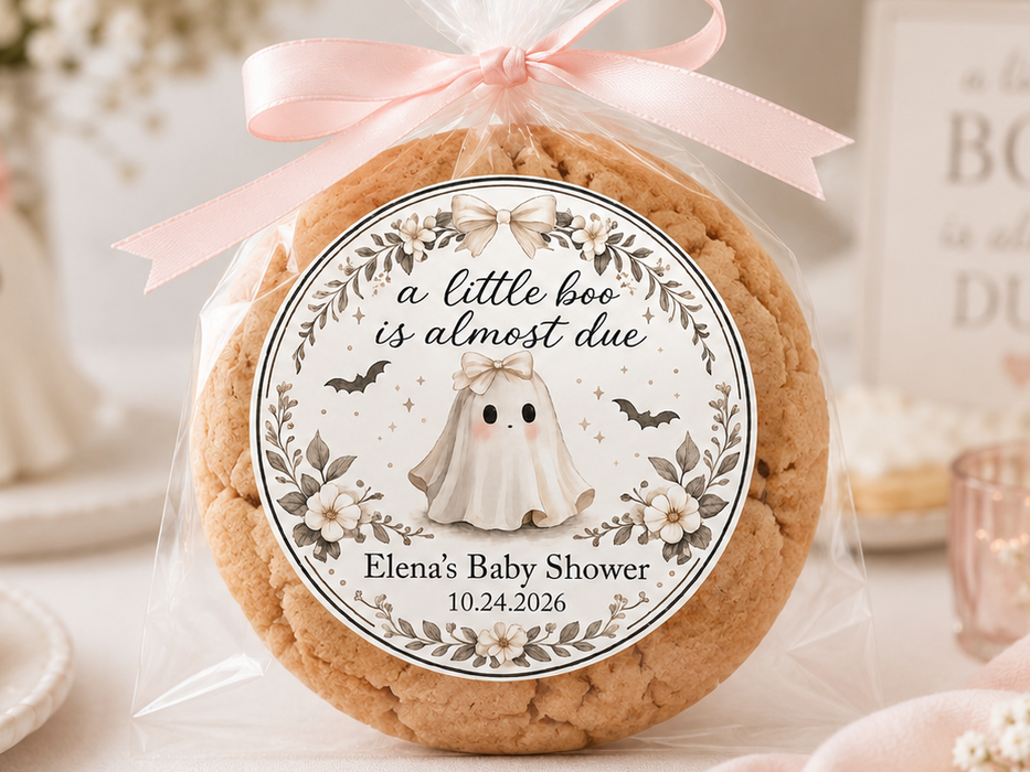 A round custom baby shower cookie wrapped in clear cellophane with a pink ribbon and a decorative printed label reading 'a little boo is almost due' and 'Elena's Baby Shower,' styled on a tabletop with pastel party decor in the background.
