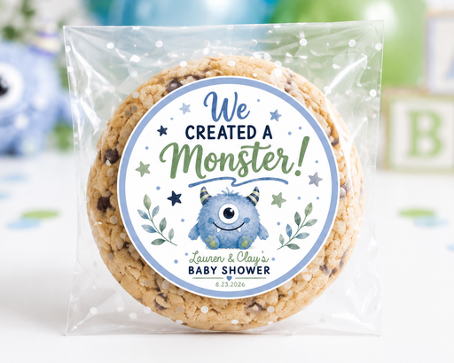A round chocolate chip cookie sealed in a clear plastic bag with a custom round label that reads 'We Created a Monster! Learn & Play's Baby Shower'. The background is a white tabletop decorated with blue and green balloons, a blue monster figurine, and wooden alphabet blocks.