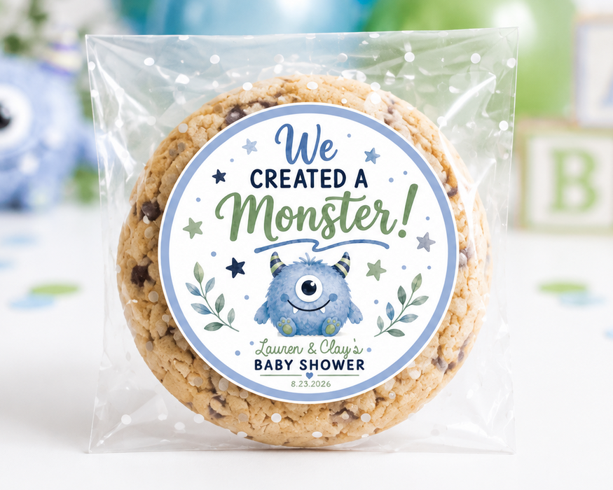 A round chocolate chip cookie sealed in a clear plastic bag with a custom round label that reads 'We Created a Monster! Learn & Play's Baby Shower'. The background is a white tabletop decorated with blue and green balloons, a blue monster figurine, and wooden alphabet blocks.