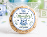 A round chocolate chip cookie sealed in a clear plastic bag with a custom round label that reads 'We Created a Monster! Learn & Play's Baby Shower'. The background is a white tabletop decorated with blue and green balloons, a blue monster figurine, and wooden alphabet blocks.