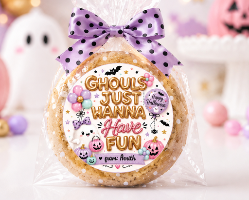 A round sugar cookie wrapped in clear cellophane and tied with a purple polka-dot bow, featuring a festive Halloween label that reads 'Ghouls just wanna Have fun' with pumpkins, ghosts, and bats. The background is a softly blurred Halloween-themed setup with pink pumpkins, pastel balloons, and a white ghost decoration.