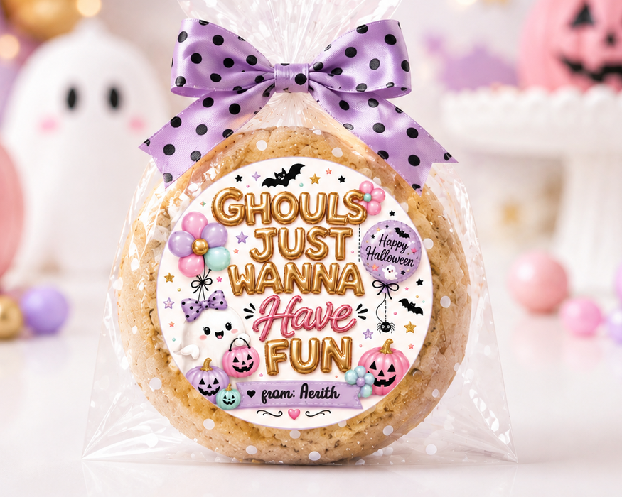 A round sugar cookie wrapped in clear cellophane and tied with a purple polka-dot bow, featuring a festive Halloween label that reads 'Ghouls just wanna Have fun' with pumpkins, ghosts, and bats. The background is a softly blurred Halloween-themed setup with pink pumpkins, pastel balloons, and a white ghost decoration.
