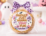 A round sugar cookie wrapped in clear cellophane and tied with a purple polka-dot bow, featuring a festive Halloween label that reads 'Ghouls just wanna Have fun' with pumpkins, ghosts, and bats. The background is a softly blurred Halloween-themed setup with pink pumpkins, pastel balloons, and a white ghost decoration.