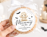 A hand holds a packaged round cookie decorated with a white ribbon and a custom round label that reads 'She's a Mummy to be' with a mummy illustration and baby shower details. The background is a softly blurred Halloween-themed setting with pumpkins, a ghost, and warm lights.