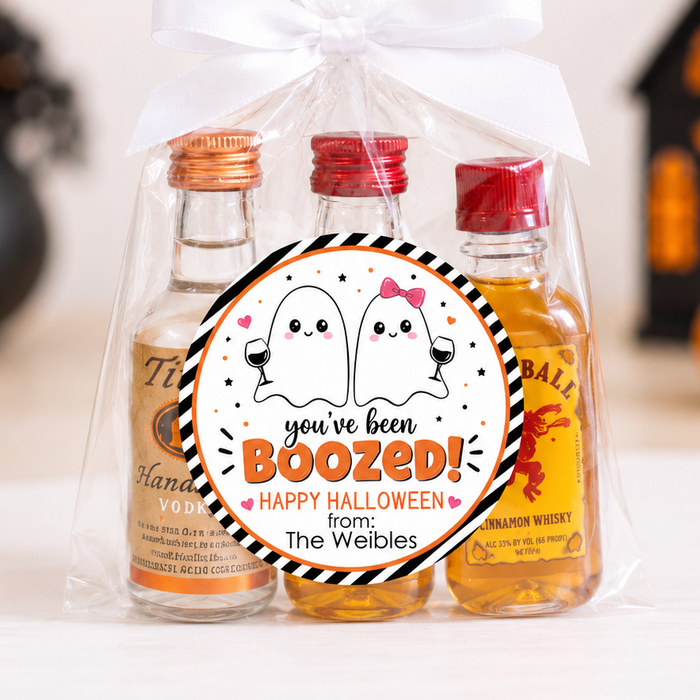 A Halloween-themed gift set in a clear cellophane bag tied with a white ribbon, containing three miniature whiskey bottles with orange and red caps. A round sticker on the front reads 'you've been BOOZED! Happy Halloween from The Weebles' with cartoon ghosts.