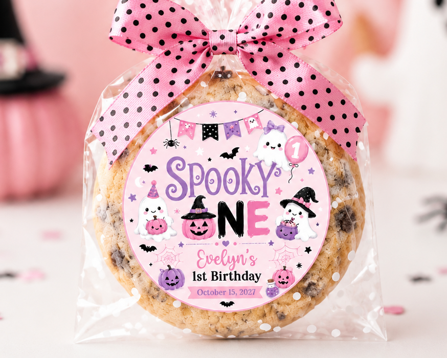 A round chocolate chip cookie wrapped in clear cellophane and tied with a pink polka-dot bow, featuring a custom 'Spooky One' birthday label. The background is a soft pink Halloween-themed setting with blurred pumpkins, a ghost, and a cupcake.