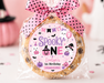 A round chocolate chip cookie wrapped in clear cellophane and tied with a pink polka-dot bow, featuring a custom 'Spooky One' birthday label. The background is a soft pink Halloween-themed setting with blurred pumpkins, a ghost, and a cupcake.