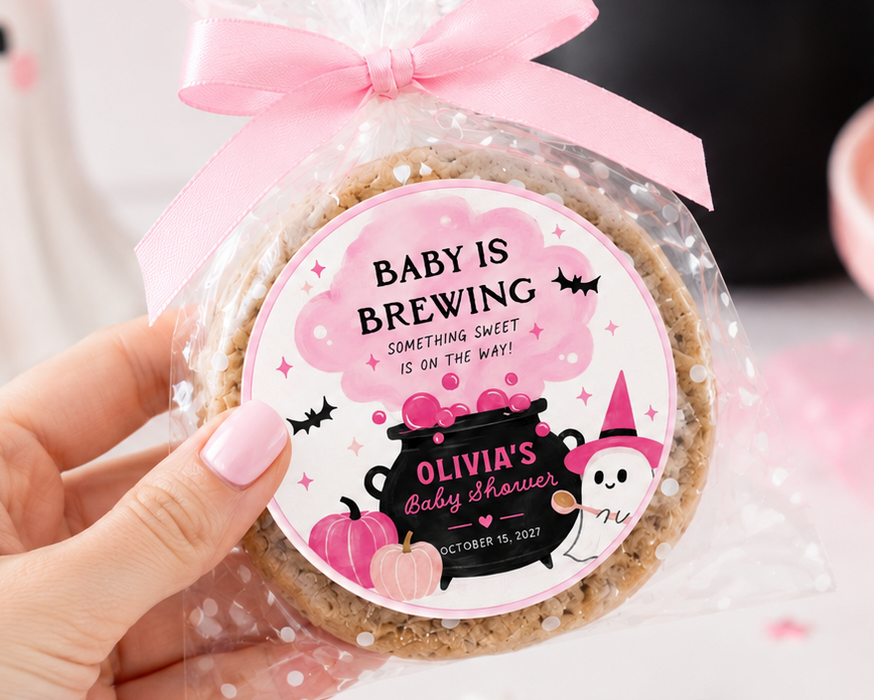A hand holds a round baked good wrapped in clear cellophane and tied with a pink ribbon, featuring a custom round label that reads 'BABY IS BREWING' with a cartoon witch and cat. The background is a styled pink Halloween-themed table setting with cupcakes, a ghost decoration, and fairy lights.